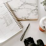 black nikon dslr camera on white notebook beside white ceramic mug